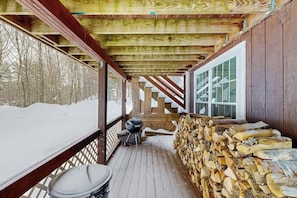 Eine Holzterrasse mit verschneitem Untergrund, einem Grill und einem Holzstoß.