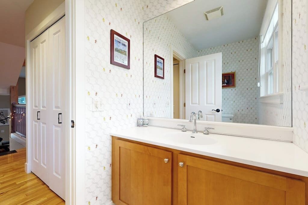 A bathroom with a white tiled wall, wooden cabinets, a large mirror, and a white countertop.