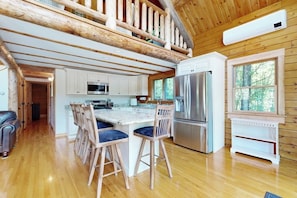 Ein Blockhauskaminareal mit einer Küche, Holzböden, einem Essbereich mit Barhocker und einem Schlafzimmer im oberen Bereich mit Holzbalkendecke.