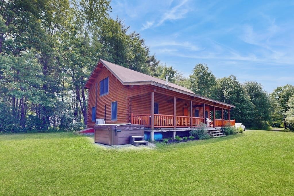 A wooden cabin with a porch, a hot tub, and a car parked nearby.