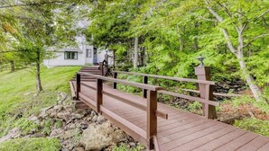 A wooden bridge leading to a white building surrounded by lush greenery.