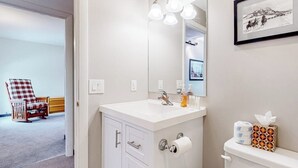 Ein Badezimmer mit einem weißen Waschbecken, einem Spiegel und einer Toilette. An der Wand hängt ein gerahmtes Bild, und ein Sessel befindet sich im angrenzenden Raum.
