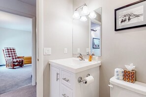 Une salle de bain avec un lavabo blanc, un miroir et des toilettes. Un tableau encadré est accroché au mur et une chaise se trouve dans la pièce adjacente.