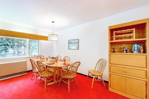 Un coin repas comprenant une table ronde, des chaises, un lustre, un radiateur, une fenêtre donnant sur des arbres et un meuble en bois avec des étagères.