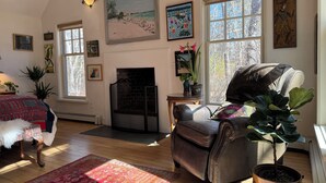 A cozy living room with a fireplace, large windows, and various artworks.