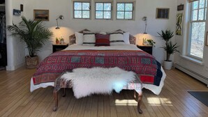 A bedroom with a large bed, two windows, a wooden floor, and a bench.