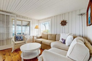 A room with a white wooden ceiling, a beige sofa, a white round table, and a wicker chair. - Charming oceanfront house with incredible beach views, BBQ area & firepit (York)