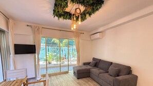 A living room with a grey sofa, wooden table, and chairs, a TV mounted on the wall, and a large sliding glass door leading to an outdoor area with plants. - Terrasse & Calme • 8 min mer • Garage privé (Antibes)