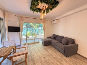 Ein Wohnzimmer mit grauem Sofa, Holztisch und Stühlen, einem an der Wand befestigten Fernseher und einer großen Schiebeglastür, die zu einem Außenbereich mit Pflanzen führt.