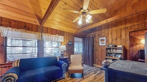 A cozy cabin interior with a wooden ceiling, a blue sofa, a chair, a fireplace, and a bookshelf. - Lakefront camp with cozy cottage-style interior and board games - kayaks welcome (Casco)