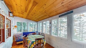 A cozy cabin interior with a wooden ceiling, a dining table with chairs, a blue sofa, and large windows offering a view of trees. - Lakefront camp with cozy cottage-style interior and board games - kayaks welcome (Casco)