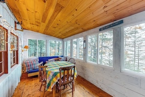 L’intérieur chaleureux d’un chalet avec un plafond en bois, une table à manger entourée de chaises, un canapé bleu et de grandes fenêtres donnant sur la forêt.