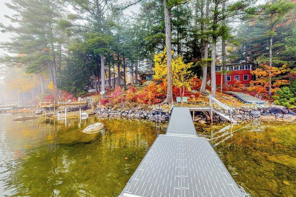 A wooden dock leading into a calm body of water, surrounded by trees with autumn foliage and a red building in the background.