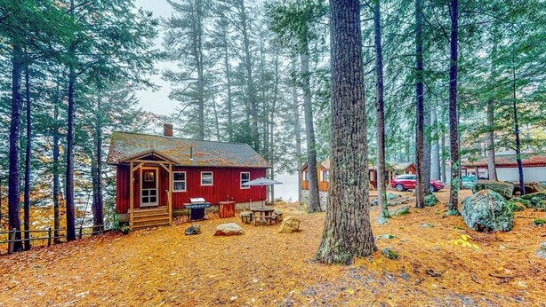 A cabin surrounded by tall trees, a wooden deck, and a covered outdoor seating area. - Lakefront camp with cozy cottage-style interior and board games - kayaks welcome (Casco)