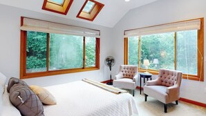 Une chambre à coucher avec un grand lit, deux fauteuils, un ventilateur sur pied et de grandes fenêtres donnant sur un paysage verdoyant.