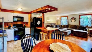 A kitchen with a microwave, stove, and refrigerator, a dining area with a wooden table and chairs, and a living room with a sofa and a coffee table. - Immaculate All-Season Retreat with Must-See Game Room! (Bethel)