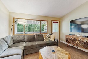 Ein Wohnzimmer mit einer Eckcouch, einem Couchtisch, einem Fernseher und einem großen Fenster mit Blick auf Bäume.
