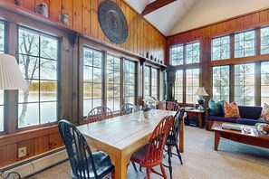A wooden interior with large windows, a dining table, chairs, a sofa, and a lamp.