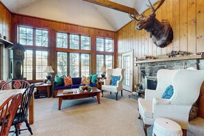 A cozy living room with a stone fireplace, wooden walls, and a mounted deer head.