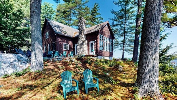 A cabin with a stone chimney, surrounded by trees and two green Adirondack chairs. - Tranquil Lakefront Retreat with large outdoor patio, beach and Stunning Views (Denmark)