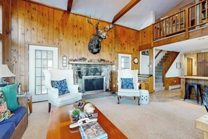 A spacious living room with a stone fireplace, wooden walls, and a mounted deer head.
