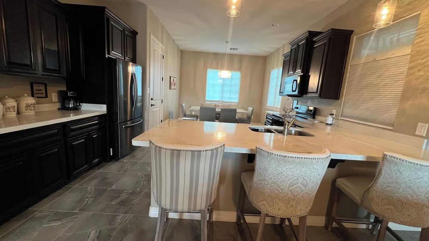A kitchen with dark cabinetry, a central island with bar stools, and a dining area in the background.