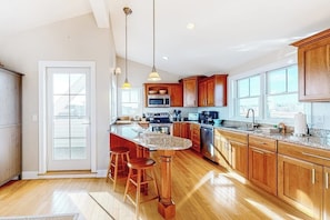 A spacious kitchen with wooden cabinets, a central island with stools, a large window, and a door leading outside. - Lovely oceanfront home with balcony, central AC, & washer/dryer (Wells)