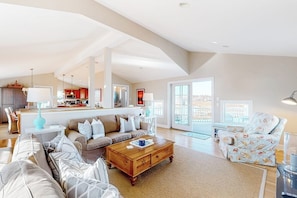 A spacious living area with a sofa, coffee table, and patterned armchair. A kitchen area is visible in the background. - Lovely oceanfront home with balcony, central AC, & washer/dryer (Wells)