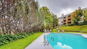 A swimming pool with a metal ladder, surrounded by a grassy area and trees, with a building in the background.