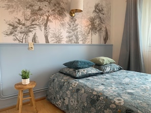 A bedroom with a floral-patterned bedspread, a wall-mounted light fixture, and a small wooden stool with a plant. - Bright Room - Centre (Lourdes)