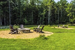 A forested outdoor area with a fire pit, chairs, and a table.