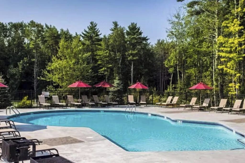 A swimming pool with lounge chairs and red umbrellas, surrounded by trees.