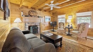 A cozy living room with a fireplace, wooden walls, a ceiling fan, and a television.
