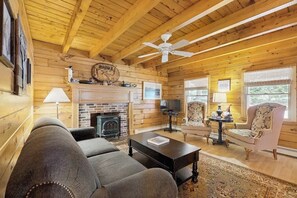 A cozy living room with a fireplace, wooden walls, a ceiling fan, and a television. - Charming Log Cabin w/ Free WiFi plus a Community Pool, Tennis, & Clubhouse (Bridgton)