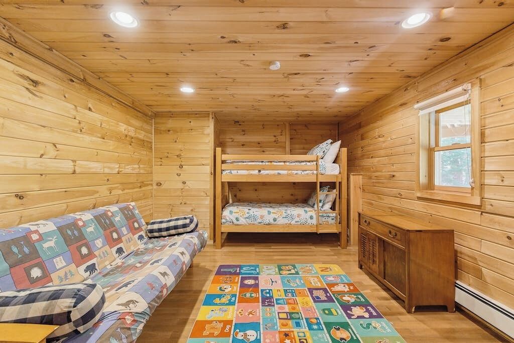 A wooden cabin interior with a bunk bed, a sofa, a colorful play mat, and a window.