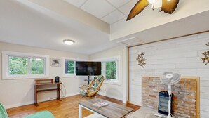 A living room with a wooden floor, a green sofa, a table with a magazine, a TV on a stand, a fireplace with a brick wall, and a ceiling fan. - Entire lakefront home on Togus Pond with decks, canoe, firepit & great view (Augusta)