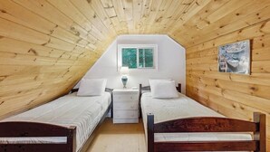 A room with wooden walls and a sloped ceiling, two beds, a window, a nightstand, and a painting.