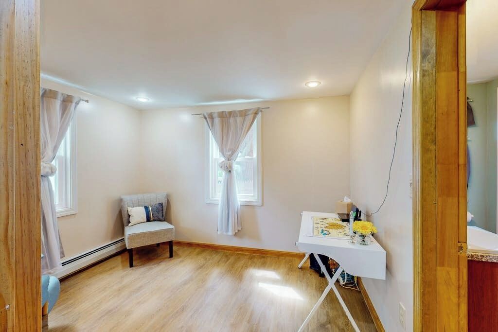 A room with a wooden floor, a white desk with a vase of flowers, a grey sofa, and two windows with curtains.