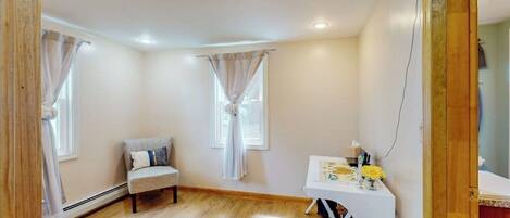A room with a wooden floor, a white desk with a vase of flowers, a grey sofa, and two windows with curtains.