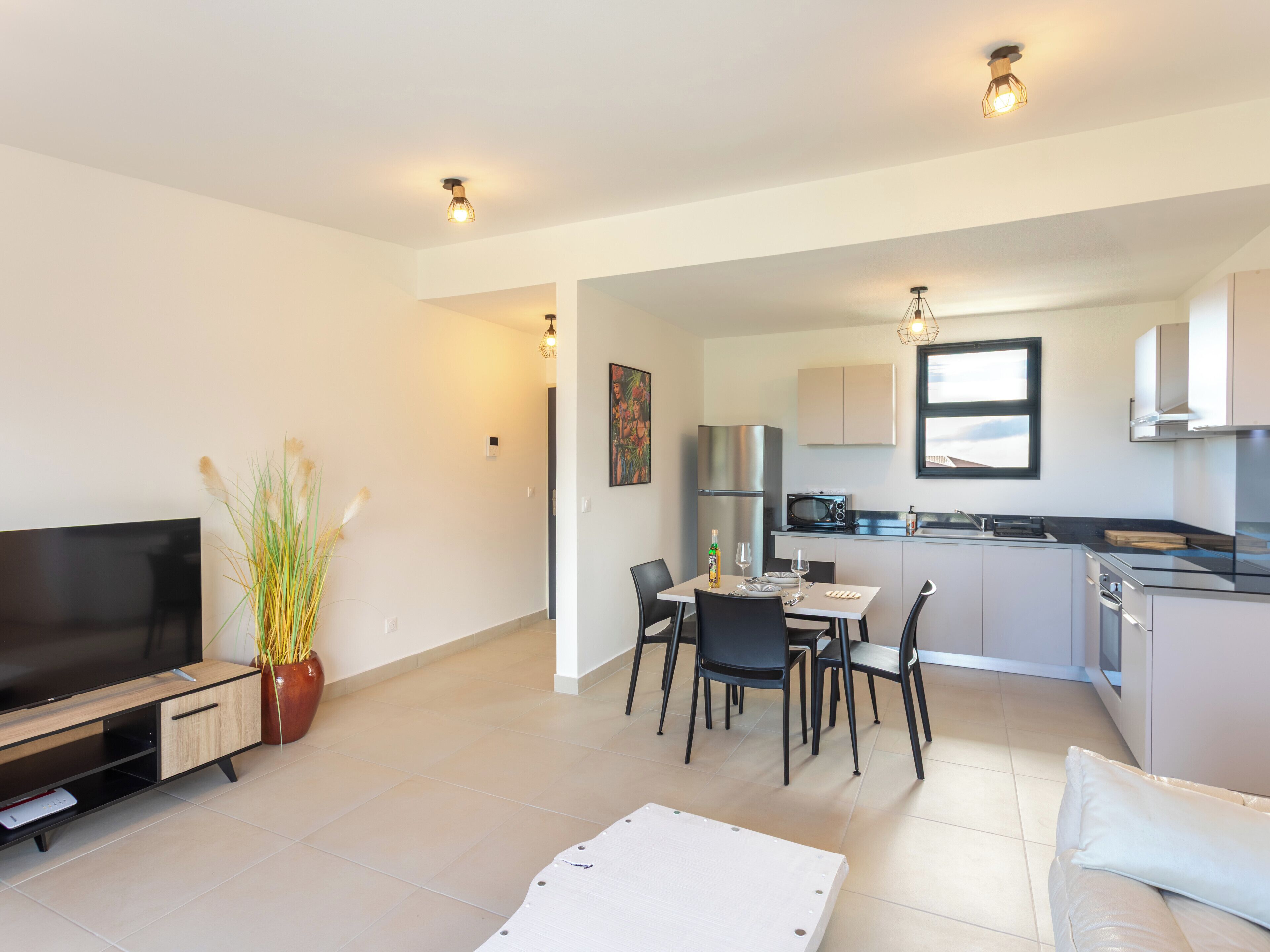 A modern living room with a flat-screen TV, a dining table with chairs, a kitchen area with cabinets and appliances, and a large window.