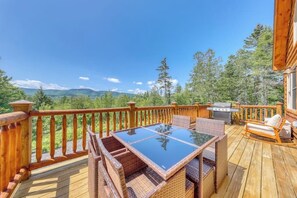 Eine Holzterrasse mit einem Tisch aus Glas und Korbstühlen, mit Blick auf eine bewaldete Landschaft.