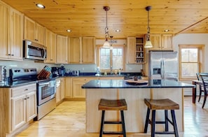 A kitchen with wooden cabinets, stainless steel appliances, a central island with stools, and pendant lighting. - The Escape at ThunderSnow Sunday River lodge with Stunning Views to Wake Up to (Bethel)