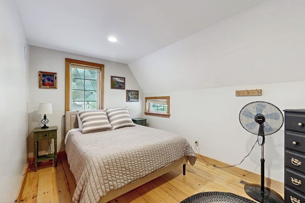 A bedroom with a bed, a standing fan, a dresser, and a window.