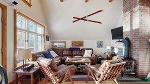 Un salon spacieux avec de hauts plafonds, une cheminée, des canapés en cuir, des fauteuils en bois et un téléviseur.