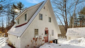 Une maison blanche, de plain-pied, avec une porte rouge, entourée de neige et d’arbres.