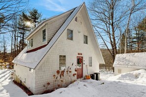 Una casa bianca a un piano con una porta rossa, circondata da neve e alberi.
