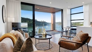 En moderne stue med sofa, lænestole, sofabord og fjernsyn. Der er en balkon med udsigt til havet og både.