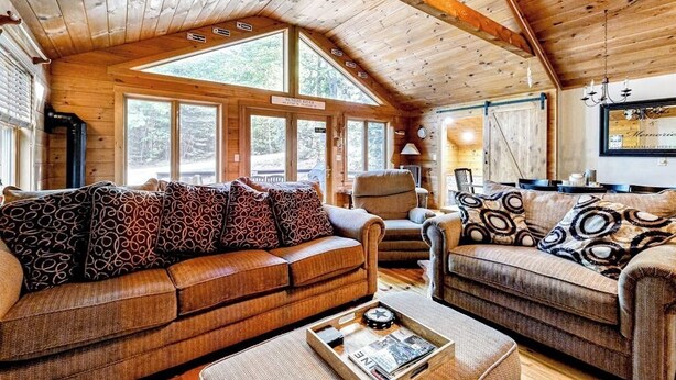 Un acogedor interior de cabaña con techo de madera, ventanas amplias, un sofá, una silla y una mesa de comedor.
