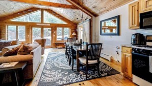 Acogedor interior de cabaña con comedor, cocina y un ventanal grande con vistas a un bosque.