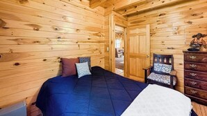 Interior de uma cabana de madeira com cama, cômoda, cadeira e porta que dá para outro quarto.